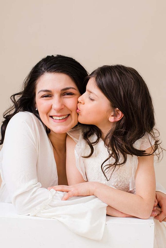 Heartwarming mother and daughter photoshoot in matching white dresses, with the young girl kissing her mother on the cheek in a London studio.