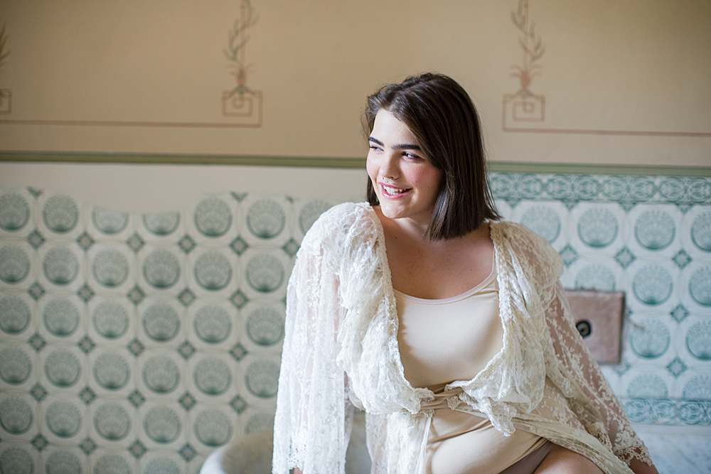 plus-size-boudoir-shoots-woman-leaning-on-bathtub-laughing-in-white-lace-gown-blue-tiles-bathroom